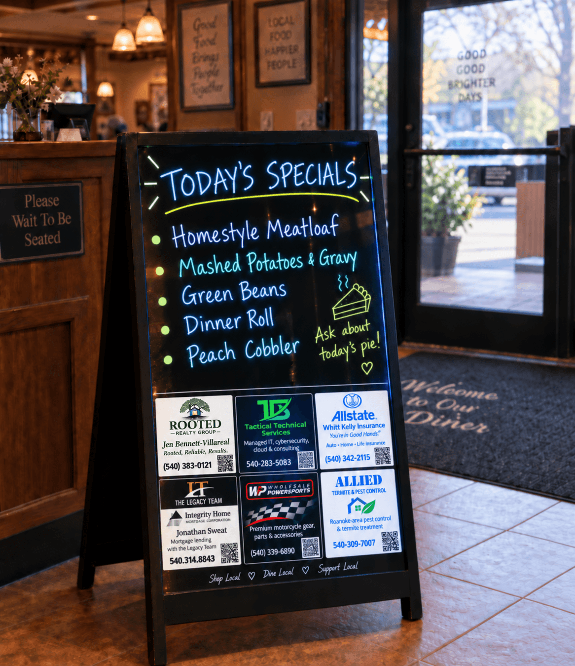 An illuminated black A-frame specials board at a restaurant entrance, with handwritten daily specials above six local business advertisements.